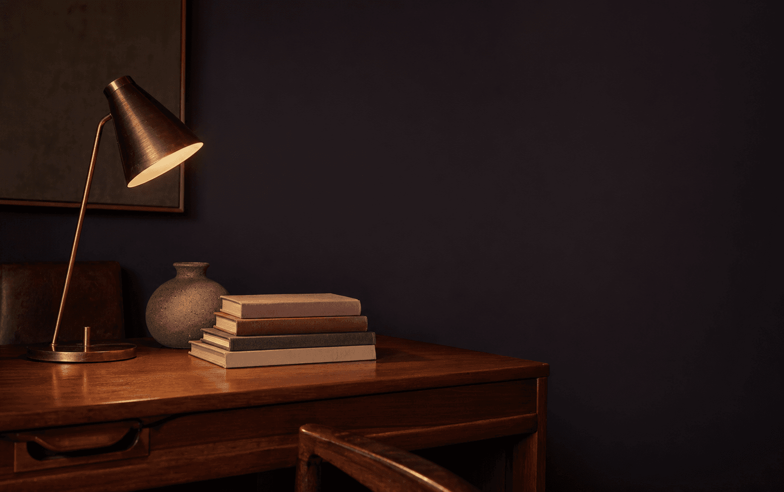 A warm walnut table with a brass lamp, stacked books, ceramic vessel, and quiet clinical workspace light.