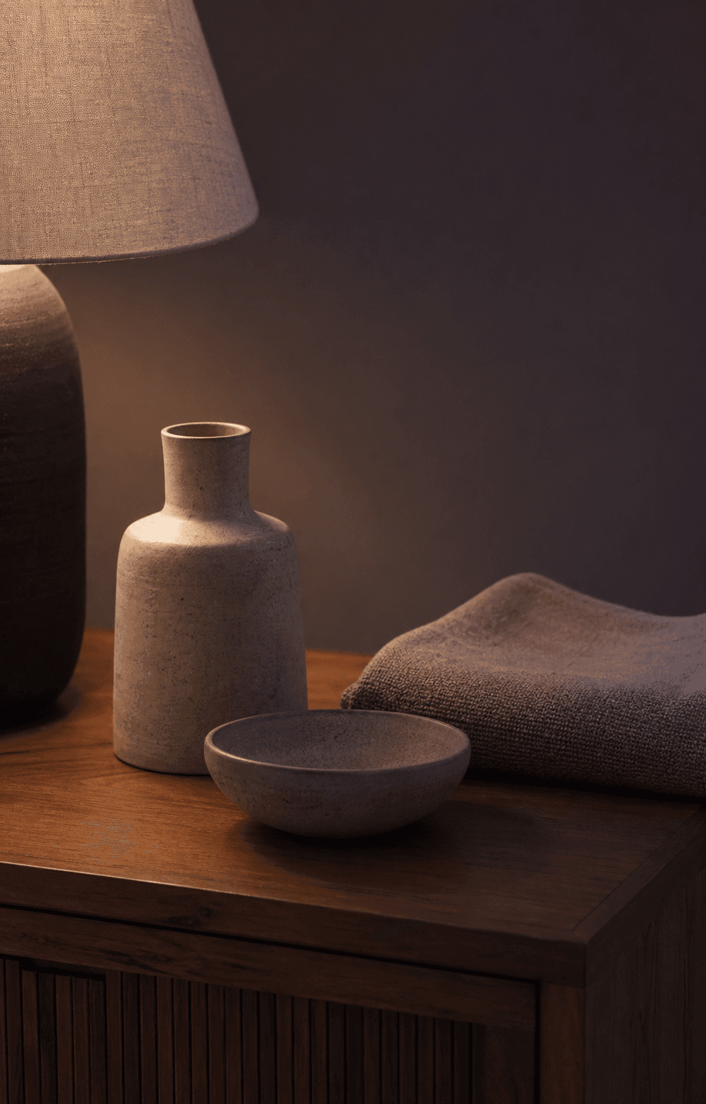 A close, quiet still life of a lamp, ceramic vessels, folded textile, and warm wood surface.